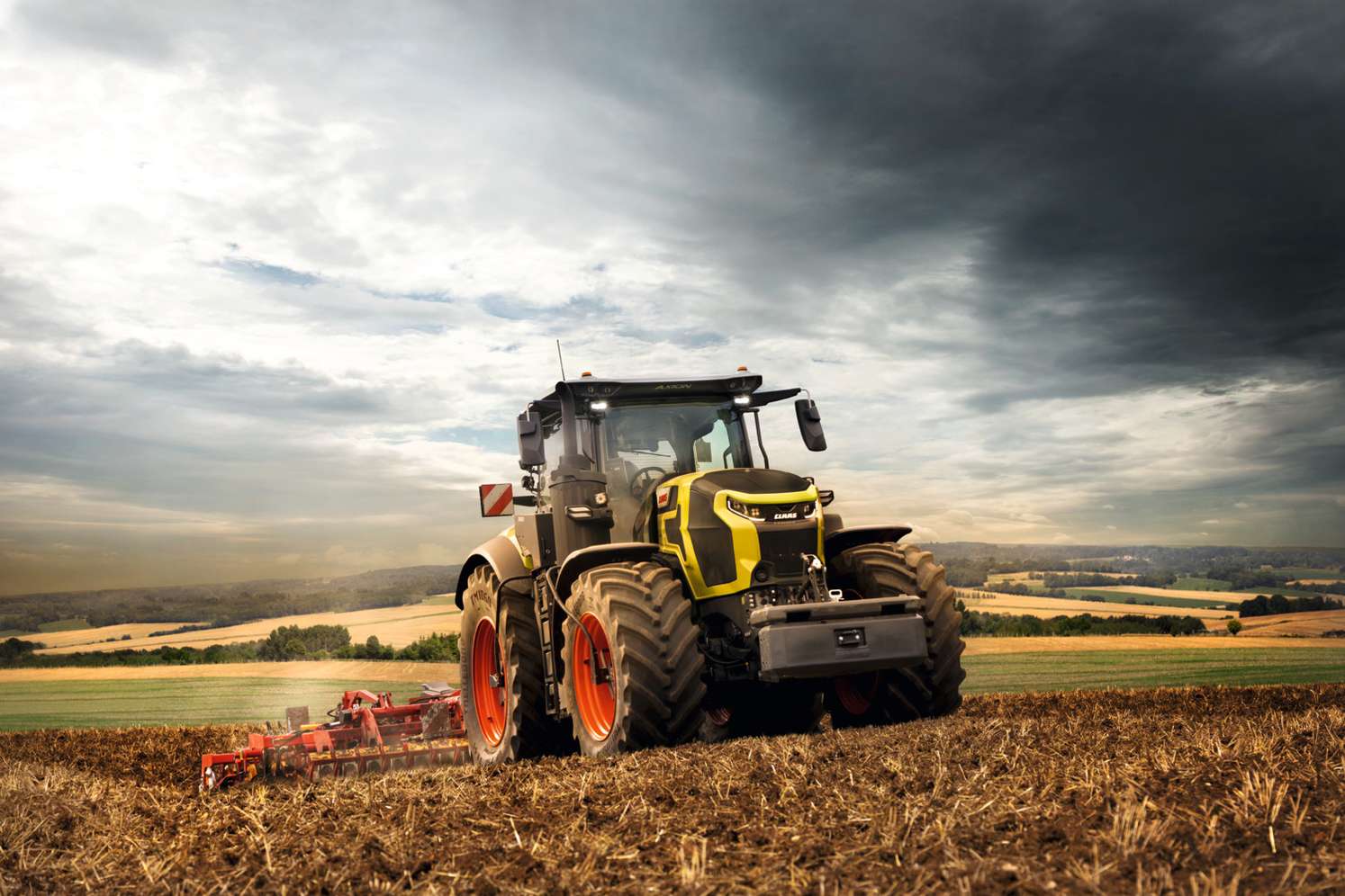 Moderner CLAAS Traktor mit gelbem Frontdesign und orangefarbenen Felgen bei der Feldbearbeitung unter dramatisch bewölktem Himmel.