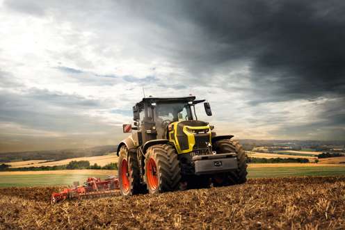 Moderner CLAAS Traktor mit gelbem Frontdesign und orangefarbenen Felgen bei der Feldbearbeitung unter dramatisch bewölktem Himmel.