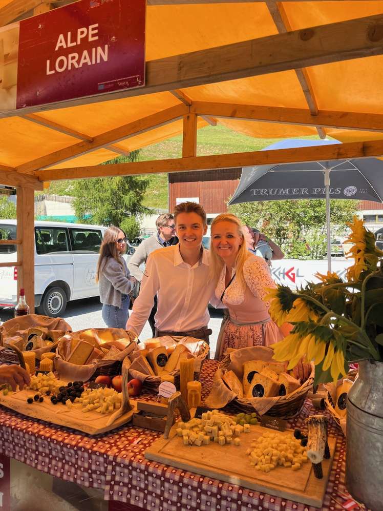 Zwei lächelnde Personen am Käsestand der Alpe Lorain auf einem Markt, mit verschiedenen Käsesorten und Sonnenblumen dekoriert.