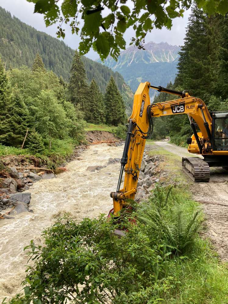 Gelber JCB-Bagger arbeitet an einem reißenden Gebirgsbach in alpiner Landschaft mit Nadelwäldern und Berggipfeln im Hintergrund.