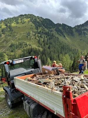Traktor mit Anhänger voller Steine und Werkzeug in den Alpen, drei Männer arbeiten, grüne Berglandschaft im Hintergrund.