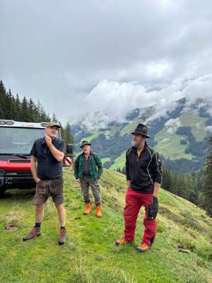 Drei Männer in Arbeitskleidung stehen auf einer Almwiese vor einem roten Aebi-Fahrzeug mit bewölkter Alpenkulisse im Hintergrund.