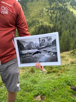 Person in roter Ortovox-Jacke hält historisches Schwarzweißfoto einer Almwirtschaft aus den 1930er Jahren vor Berglandschaft.