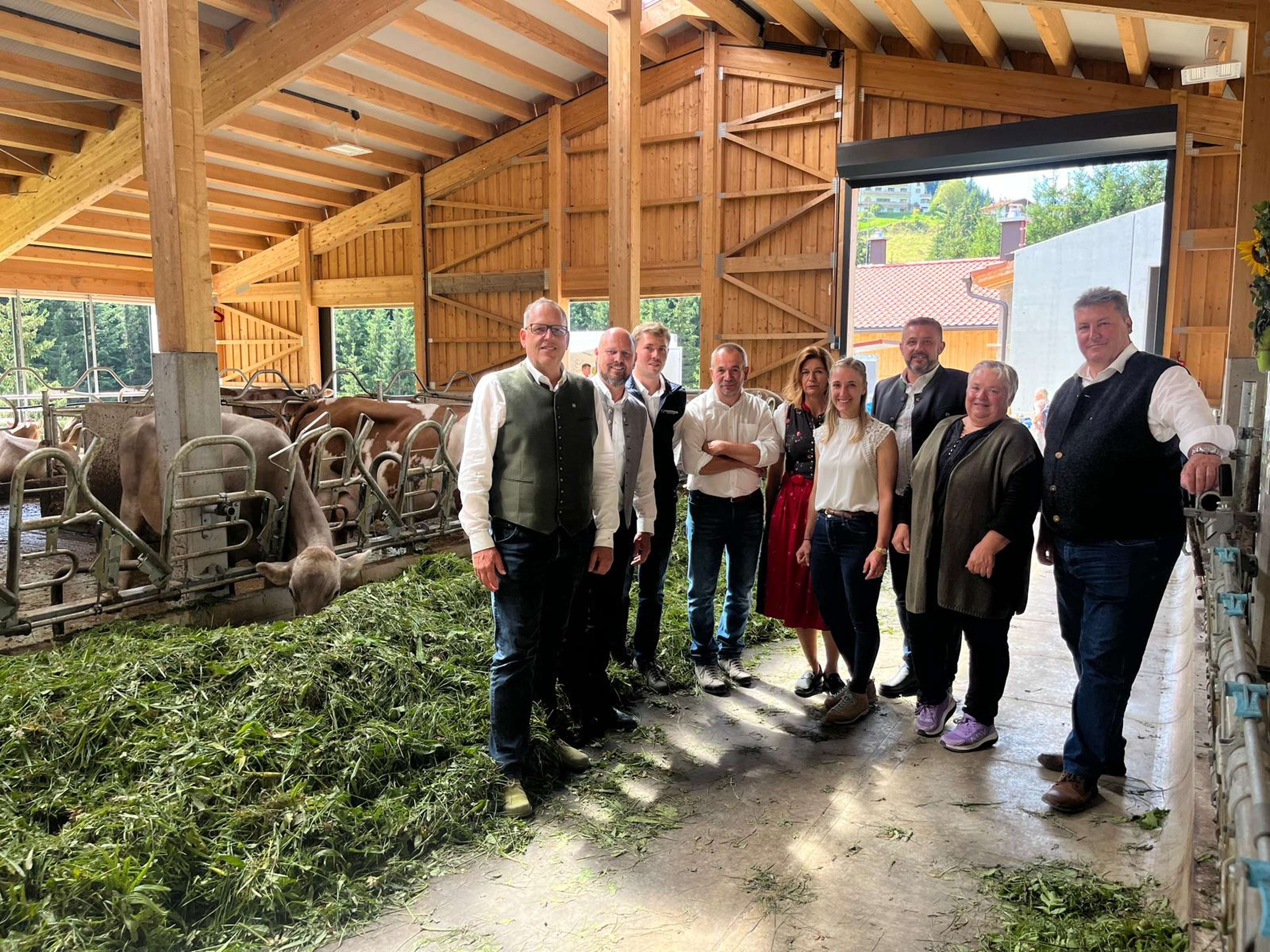 Gruppe von Menschen in einem modernen Holzstall mit Kühen, die frisches Gras fressen, bei einem Hofbesuch.