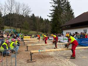 Teilnehmer schneiden Baumstämme mit Motorsägen bei einem Wettbewerb, während Zuschauer am Rand zuschauen. Im Hintergrund sind Bäume.