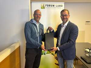 Two men in a conference room pose with a gift bag in front of a "FORUM LAND" banner featuring a plant image.