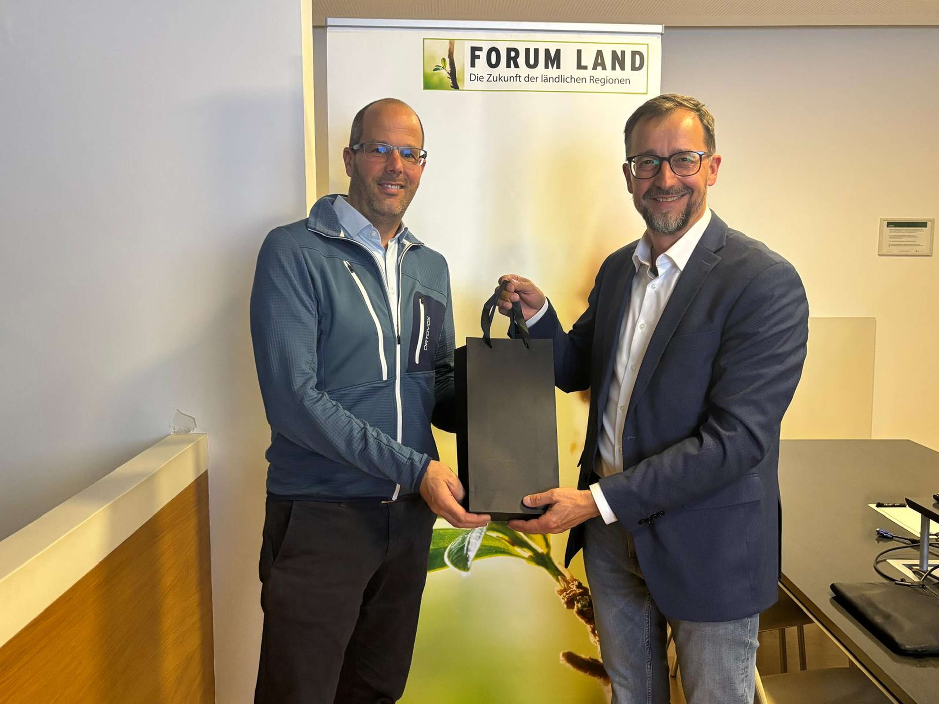 Two men in a conference room pose with a gift bag in front of a "FORUM LAND" banner featuring a plant image.