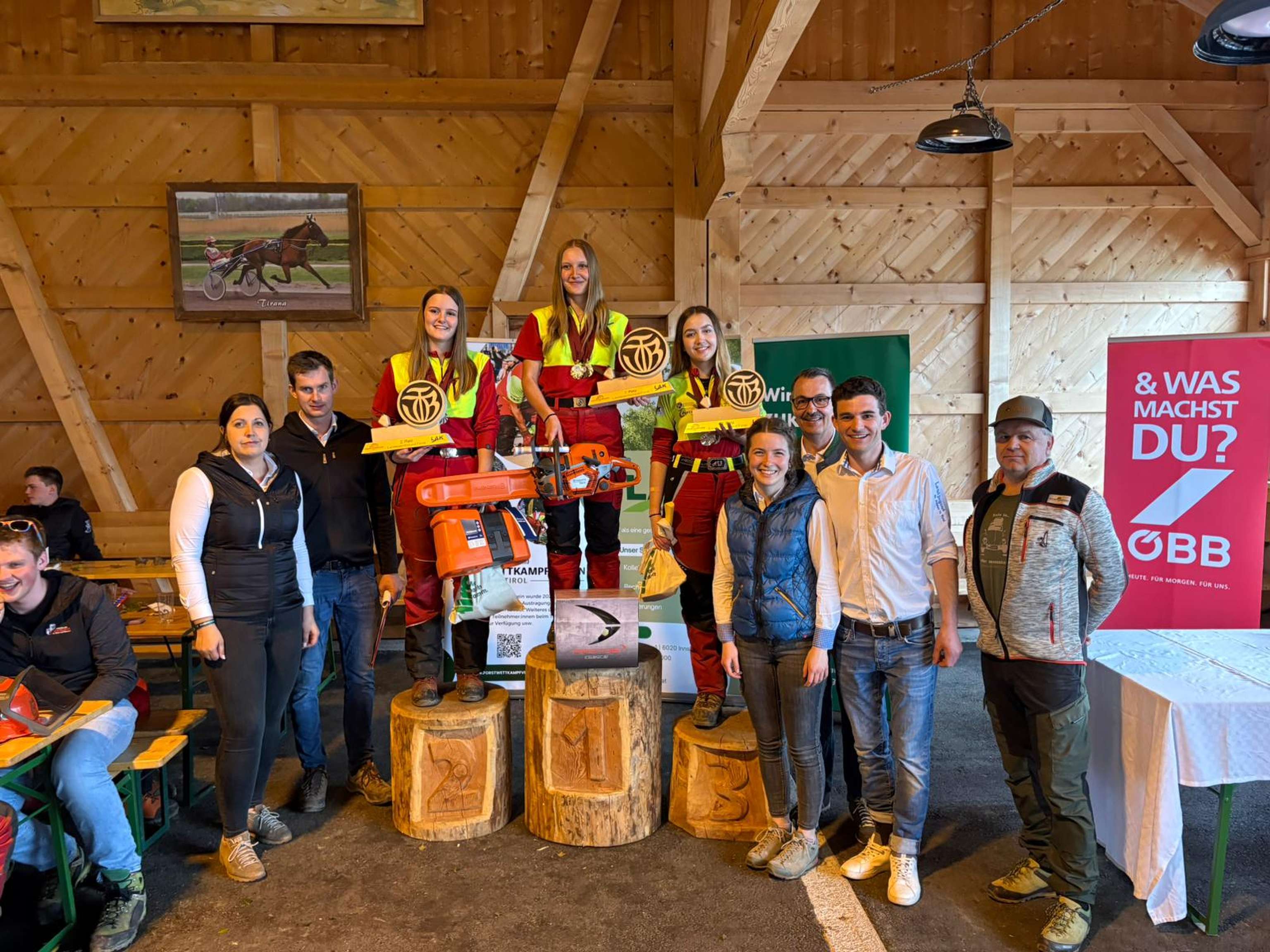 Eine Gruppe von Menschen posiert bei einer Preisverleihung in einer Holzhütte. Siegerinnen stehen auf Podesten mit Motorsägen.
