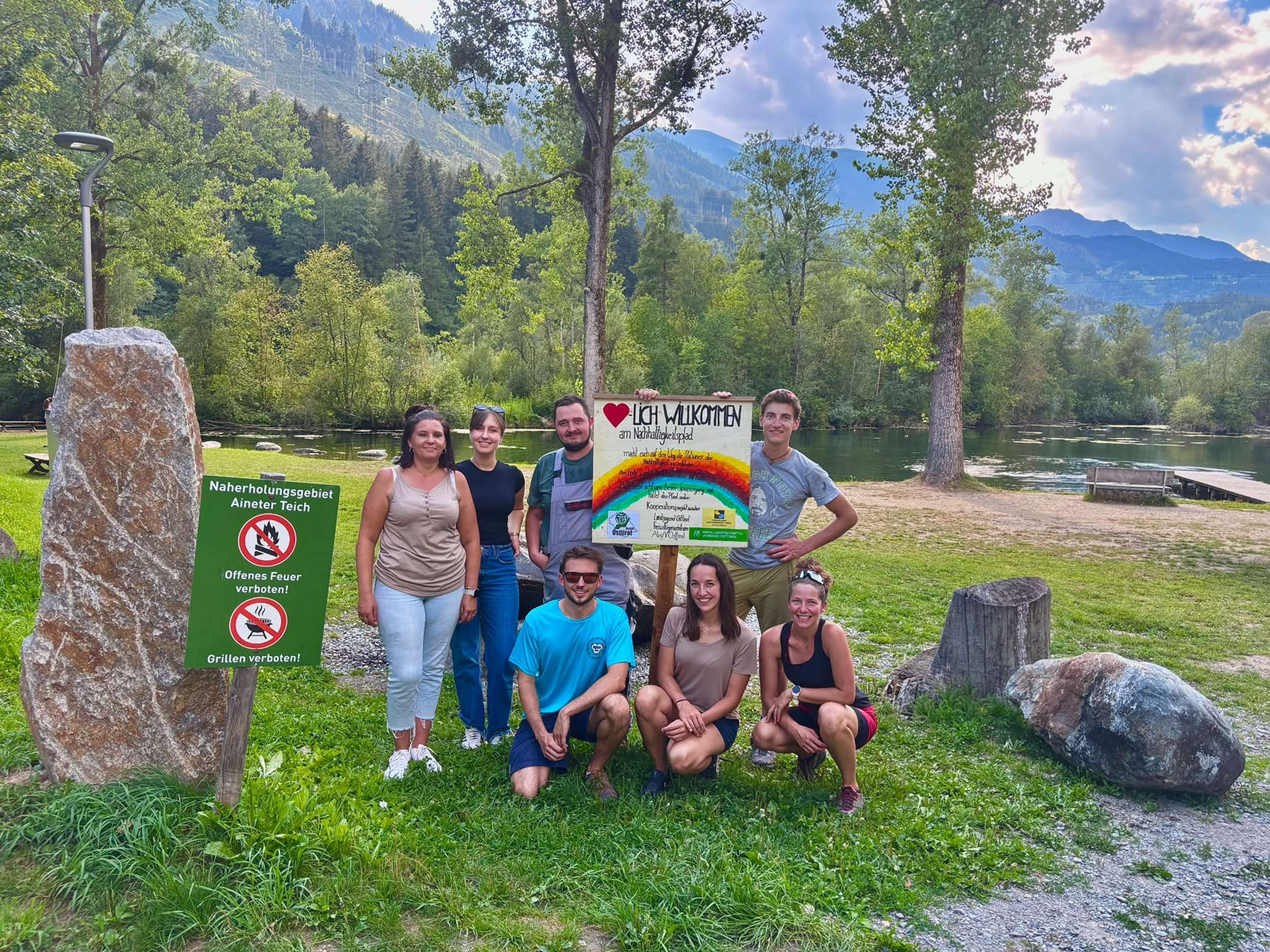 Gruppe von sieben Personen am Aineter Teich mit Willkommensschild zum Nachhaltigkeitspfad, umgeben von Bergen und Bäumen.