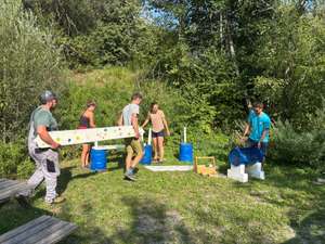Gruppe junger Menschen baut im Freien eine bemalte Bank mit blauen Fässern auf einer grünen Wiese im Wald auf.