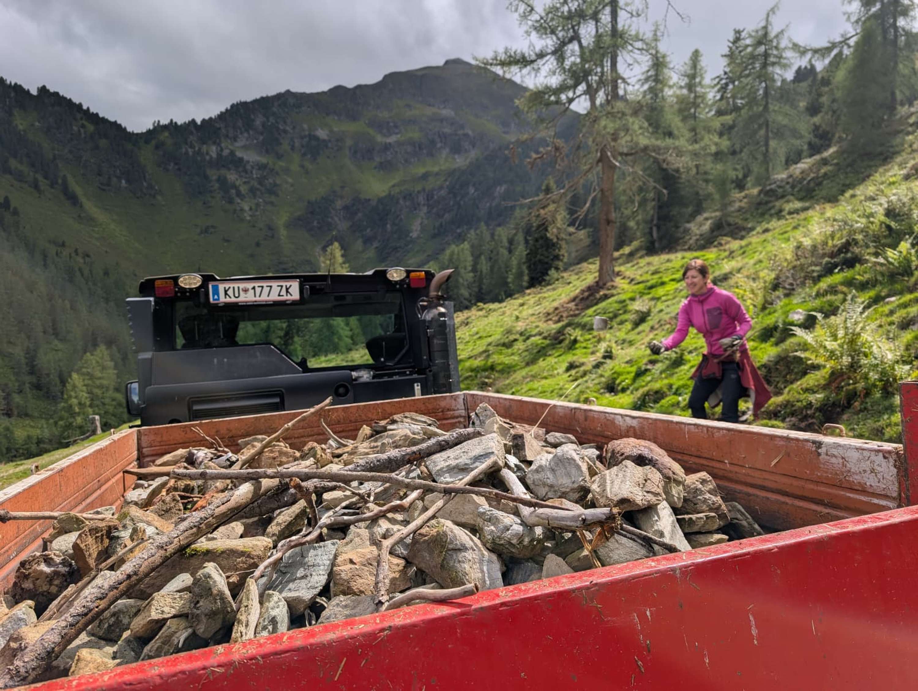 Frau in pinker Jacke arbeitet neben einem Fahrzeug mit rotem Anhänger voller Steine in alpiner Berglandschaft.