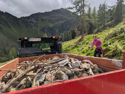 Frau in pinker Jacke arbeitet neben einem Fahrzeug mit rotem Anhänger voller Steine in alpiner Berglandschaft.