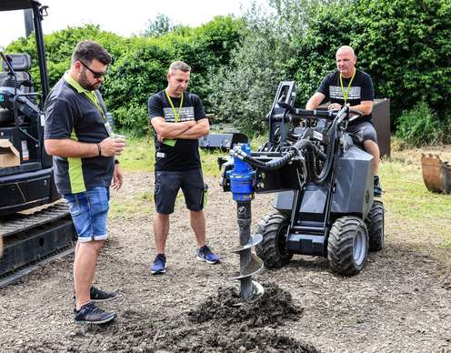 Drei Männer beobachten einen Bohrer an einem Raupenfahrzeug, das in einem grünen, bewaldeten Gebiet arbeitet.