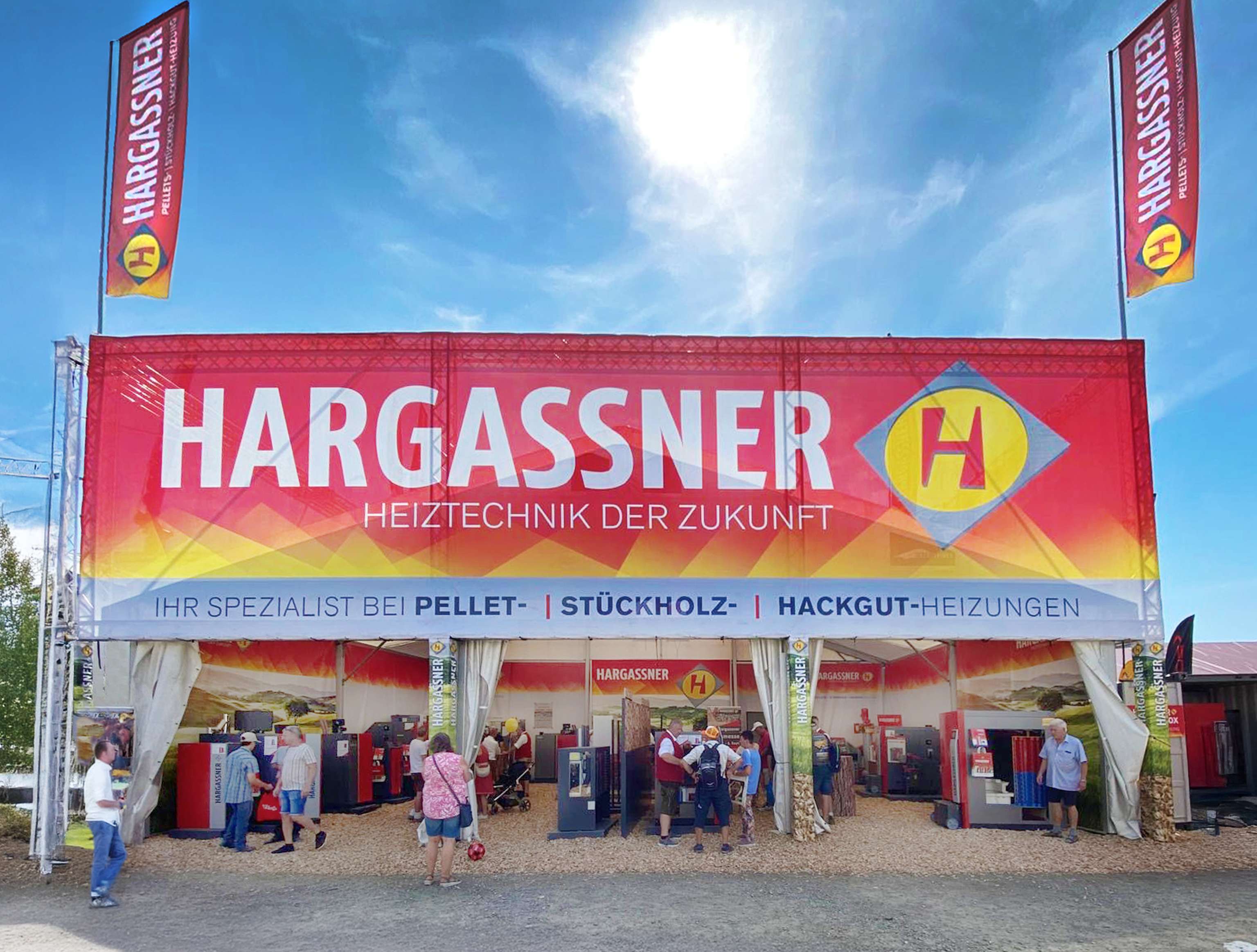 Hargassner Messestand mit rotem Banner „Heiztechnik der Zukunft", Besucher besichtigen Pellet- und Hackgut-Heizungen unter Zelt bei Sonnenschein.