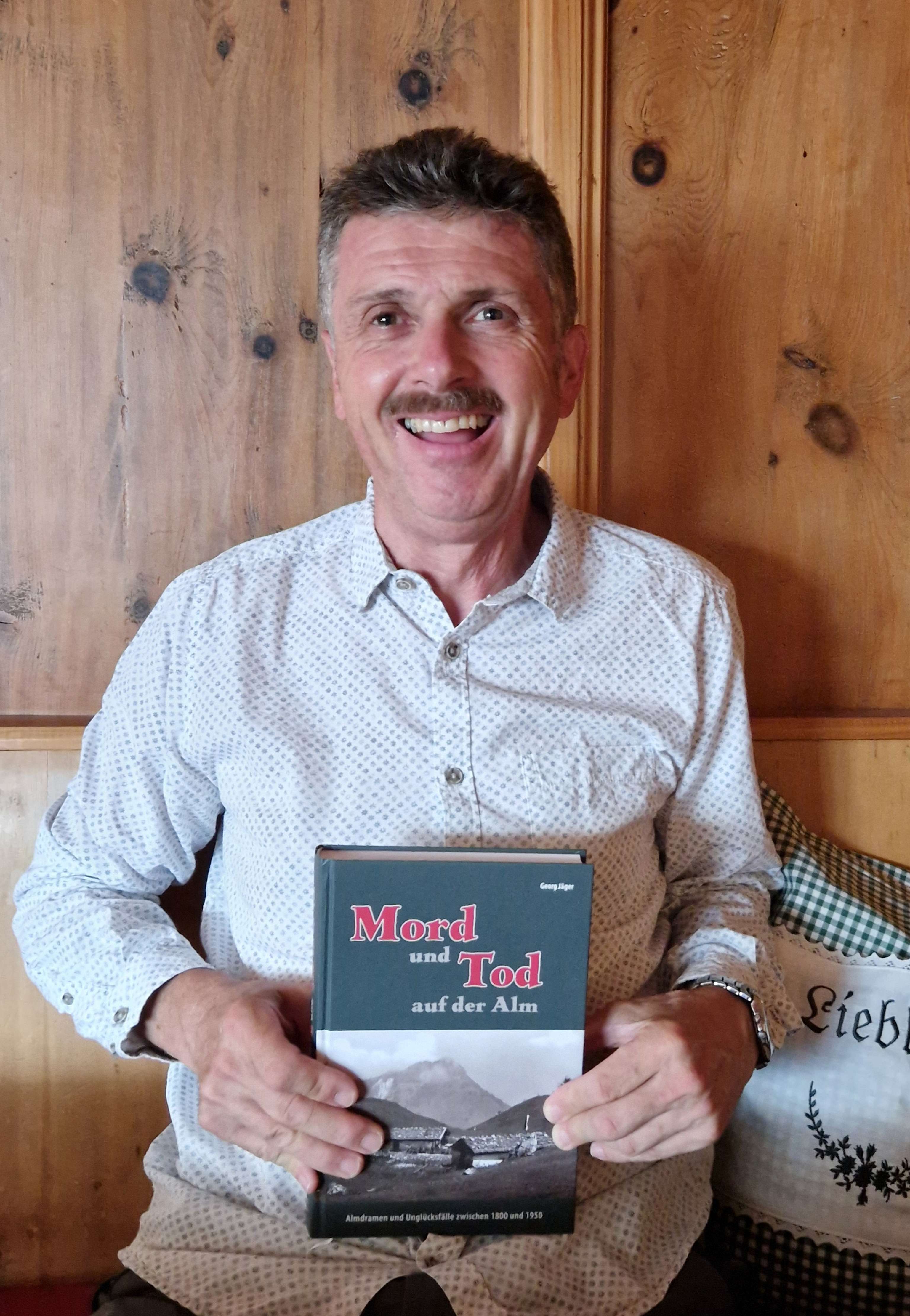 Mann mit Schnurrbart hält lächelnd das Buch „Mord und Tod auf der Alm" von Georg Jäger vor einer Holzwand.
