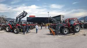 Viele rote Traktoren vor einem modernen Gebäude. Menschen betrachten die Maschinen unter einem blauen Himmel.