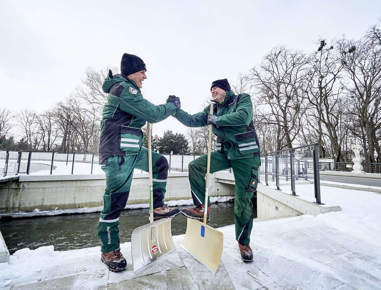 Zwei Männer in grüner Arbeitskleidung geben sich lachend die Hand, mit Schneeschiebern im Winter im Freien.