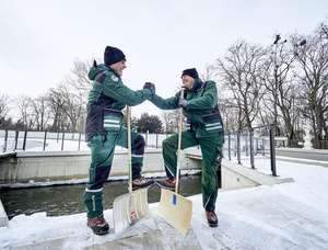 Zwei Männer in grüner Arbeitskleidung geben sich lachend die Hand, mit Schneeschiebern im Winter im Freien.