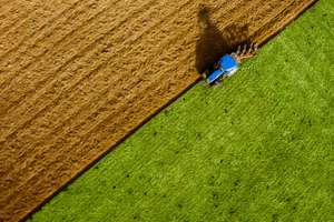 Aerial view of a blue tractor plowing a field, dividing the brown tilled soil from the vibrant green grass.