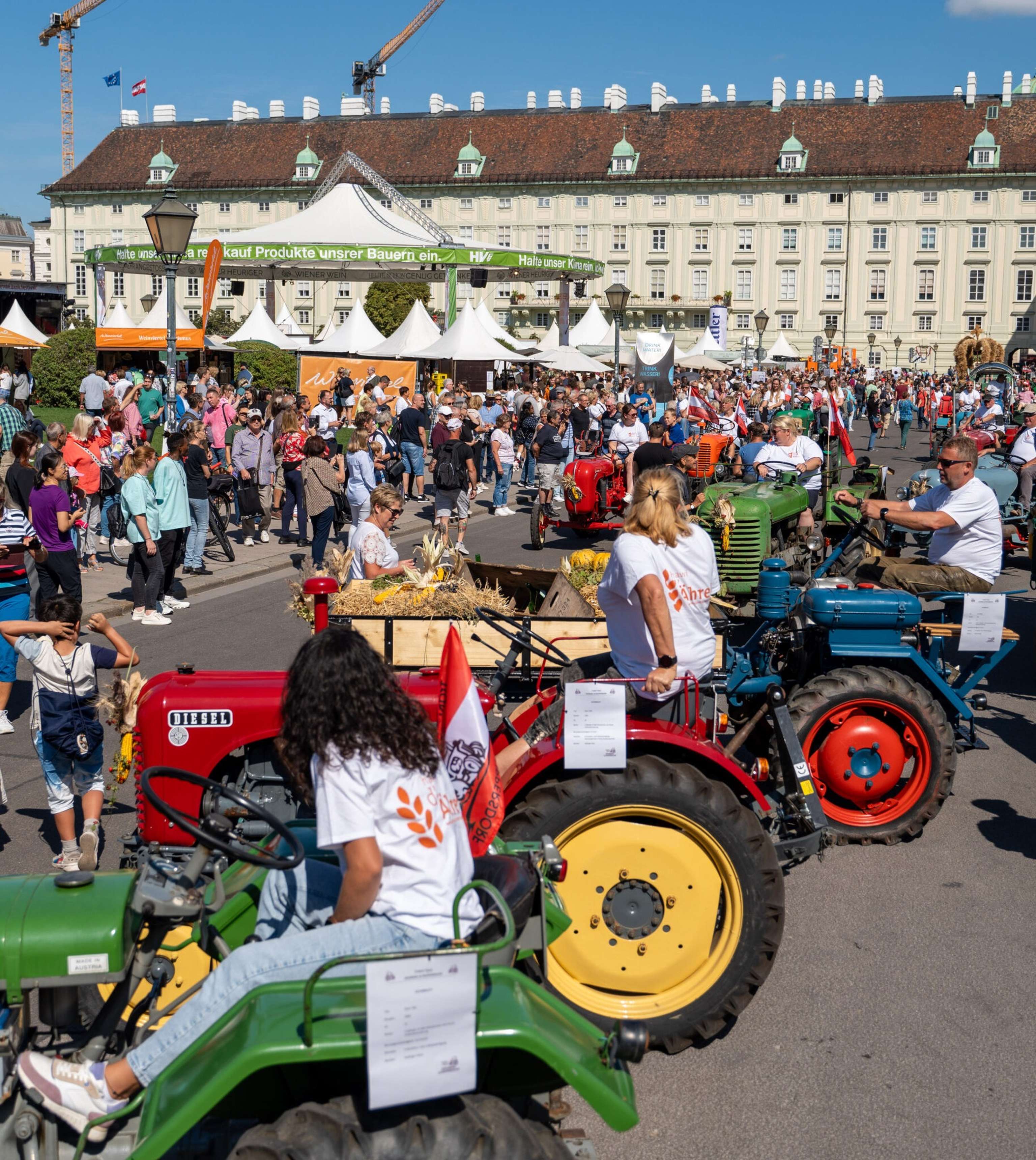 Traktorparade auf einem belebten Stadtplatz in Wien mit historischen Traktoren, Menschenmassen und weißen Festzelten bei sonnigem Wetter.