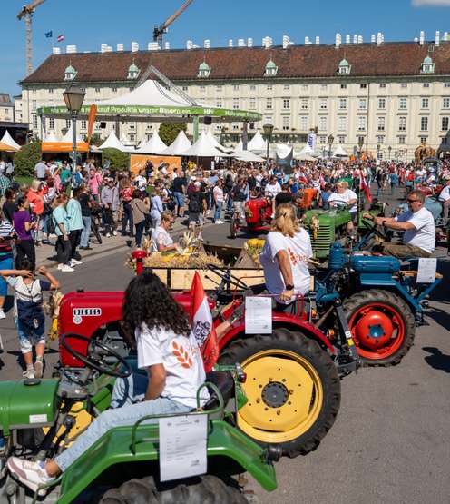 Traktorparade auf einem belebten Stadtplatz in Wien mit historischen Traktoren, Menschenmassen und weißen Festzelten bei sonnigem Wetter.