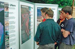 Four people stand closely examining informational display boards outdoors. The boards contain text and images about environmental topics.