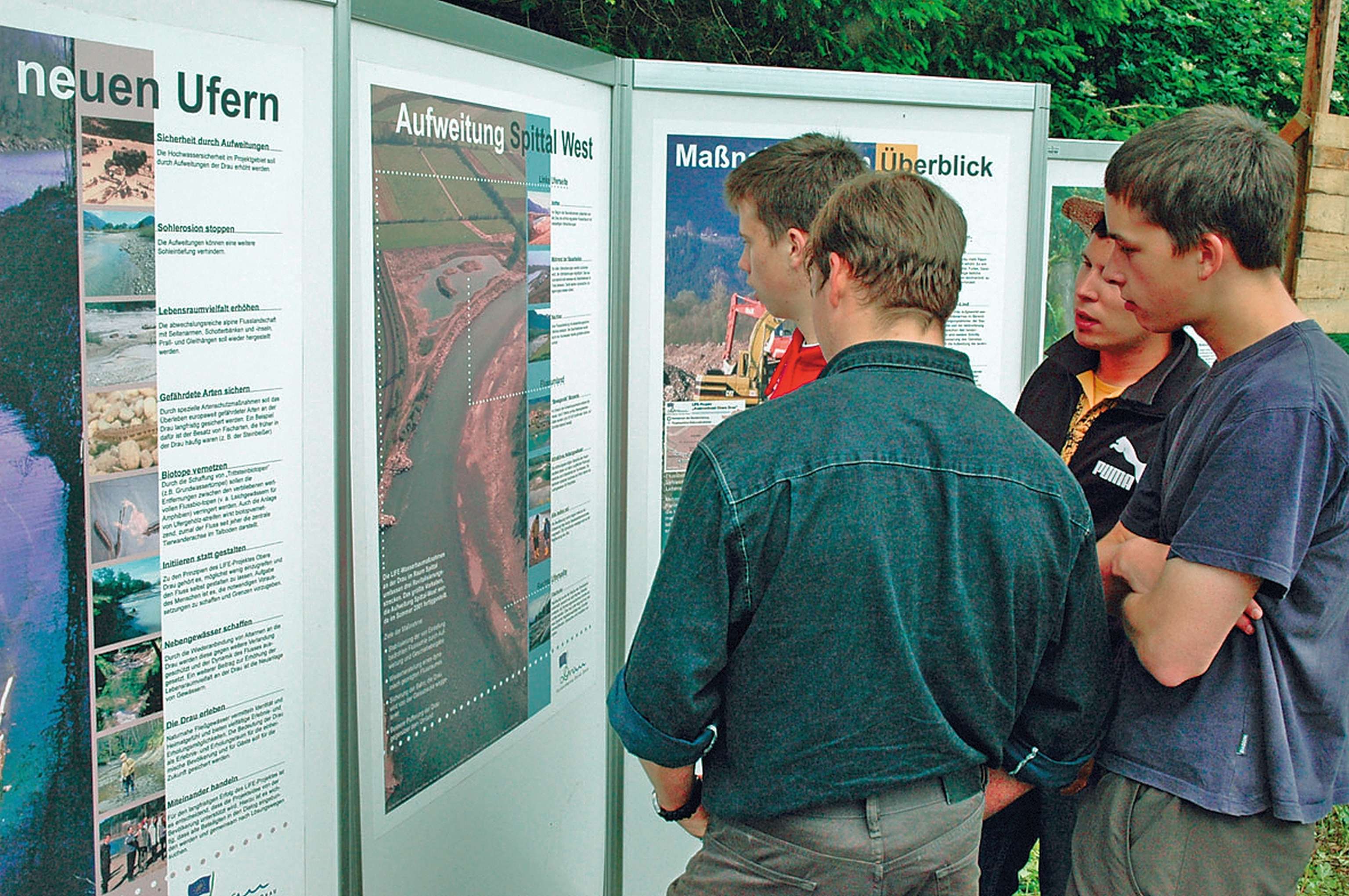 Four people stand closely examining informational display boards outdoors. The boards contain text and images about environmental topics.
