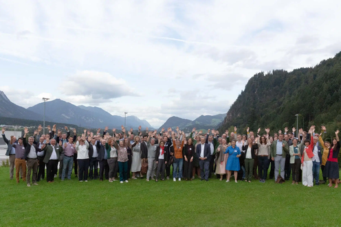 A large group of people stands on a grassy field with mountains in the background, smiling and raising their hands in celebration.