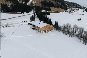 Einsames Holzgebäude in schneebedeckter Berglandschaft mit umliegendem Wald und entfernten Häusern im Hintergrund.