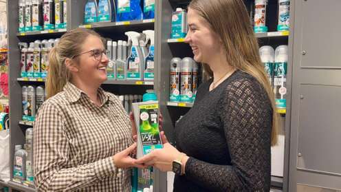Two women smiling at each other in a store aisle, holding a green product together. Shelves stocked with various cleaning supplies are in the background.