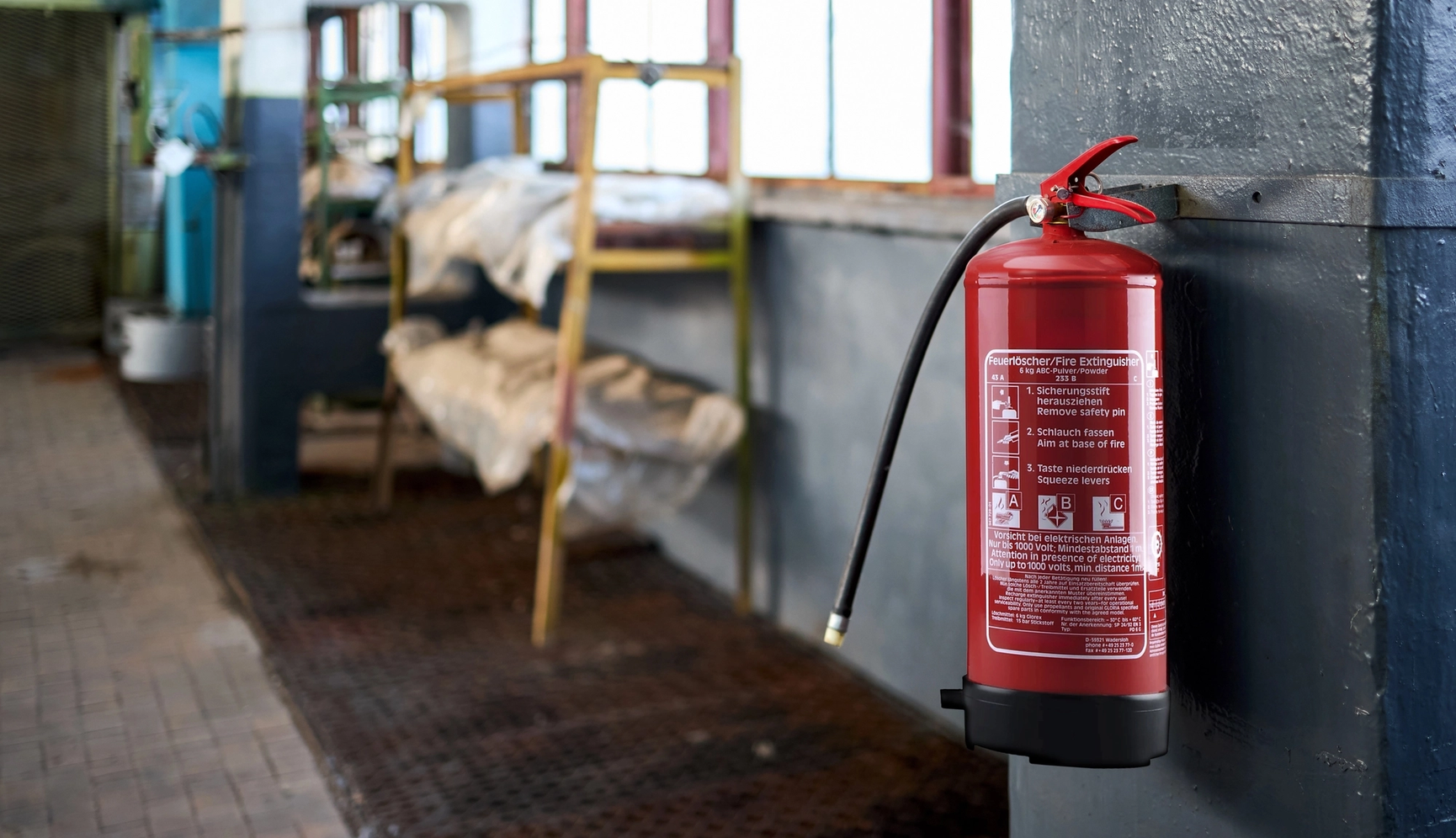 Roter Feuerlöscher an einer Wand in einem Industriegebäude, mit Anweisungen zur Bedienung aufgedruckt. Im Hintergrund unscharfe Lagerregale.