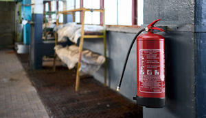Roter Feuerlöscher an einer Wand in einem Industriegebäude, mit Anweisungen zur Bedienung aufgedruckt. Im Hintergrund unscharfe Lagerregale.