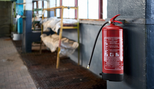 Roter Feuerlöscher an einer Wand in einem Industriegebäude, mit Anweisungen zur Bedienung aufgedruckt. Im Hintergrund unscharfe Lagerregale.
