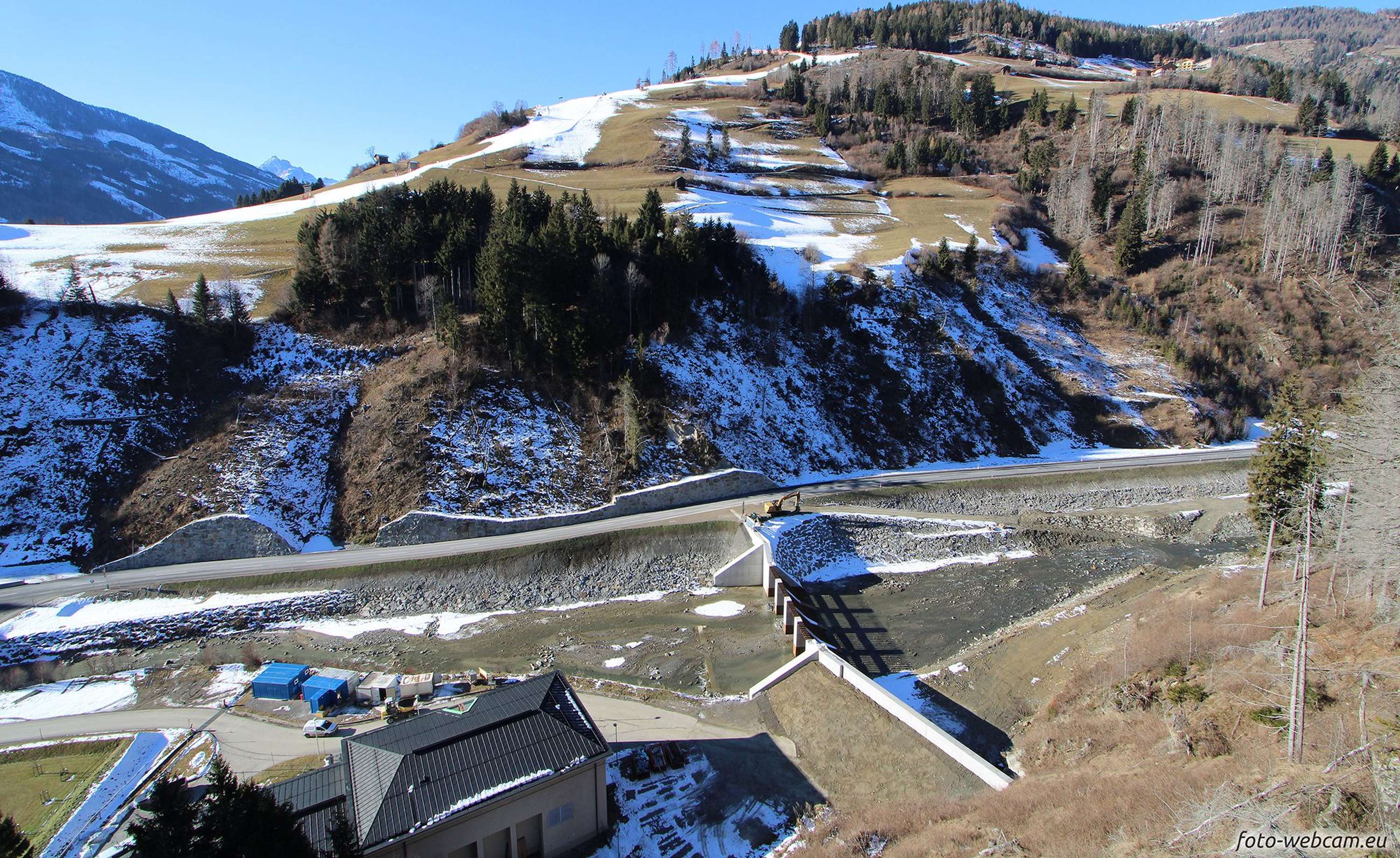 Blick auf eine alpine Landschaft mit schneebedeckten Hängen, Bäumen und einem Damm. Ein blaues Gebäude im Vordergrund.