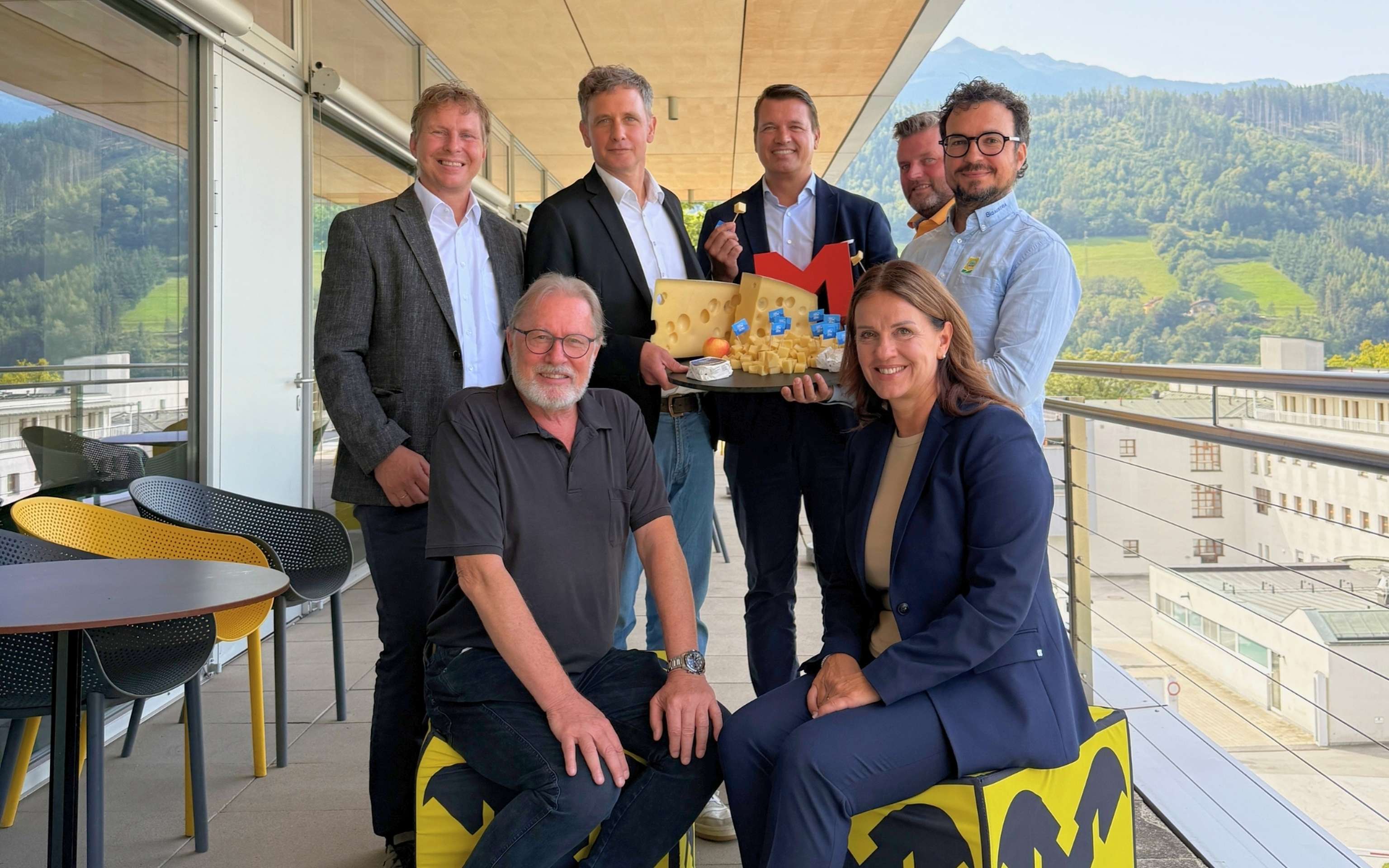 Gruppe von sieben Personen auf einer Dachterrasse mit Bergpanorama, einer hält ein Tablett mit Käse und einem roten M-Logo.