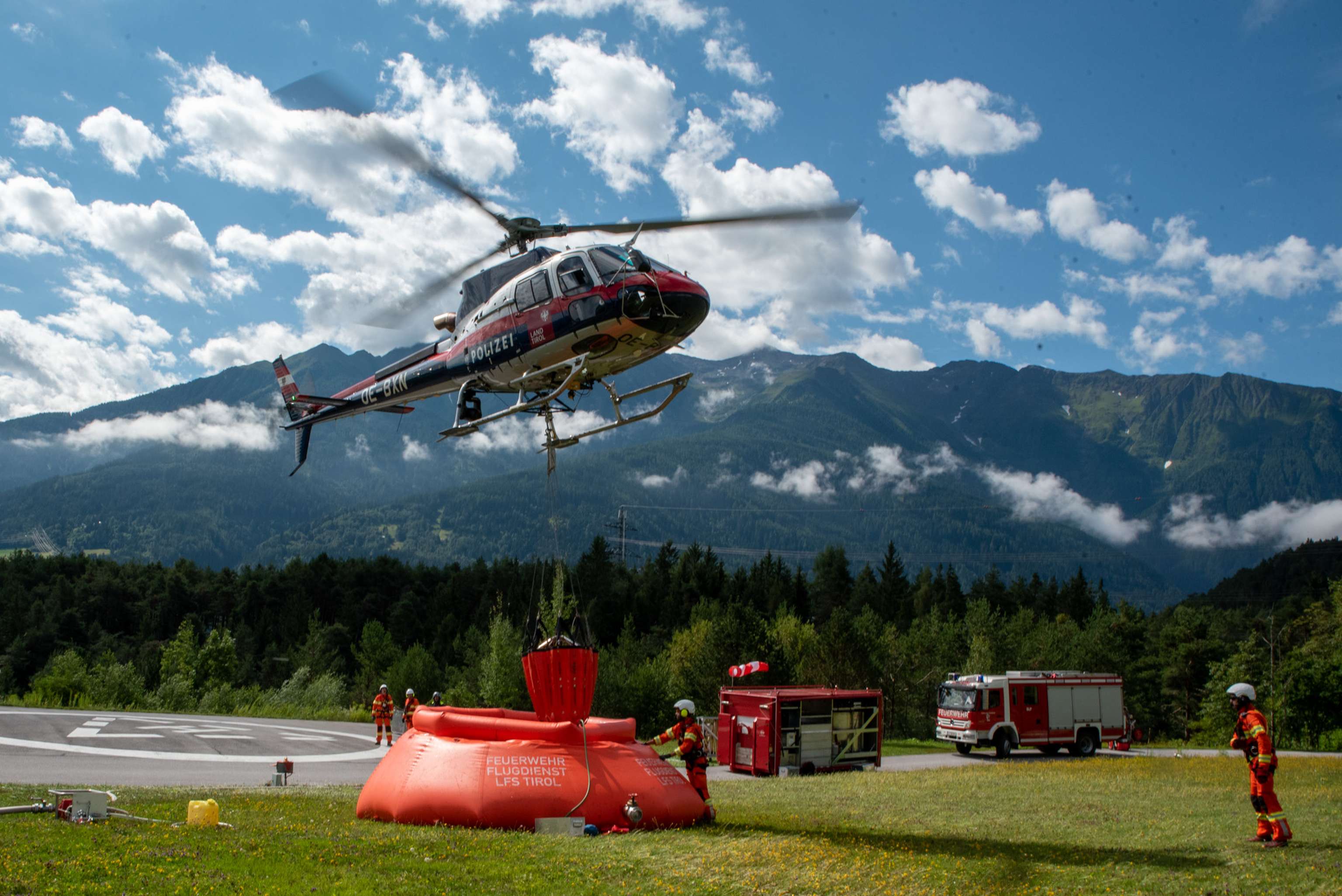 Österreichischer Polizeihubschrauber befüllt Löschwasserbehälter der Feuerwehr Tirol bei Übung in alpiner Landschaft.