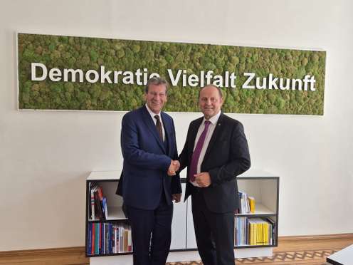 Zwei Männer im Anzug geben sich die Hand vor einem Moosbild mit der Aufschrift „Demokratie Vielfalt Zukunft".