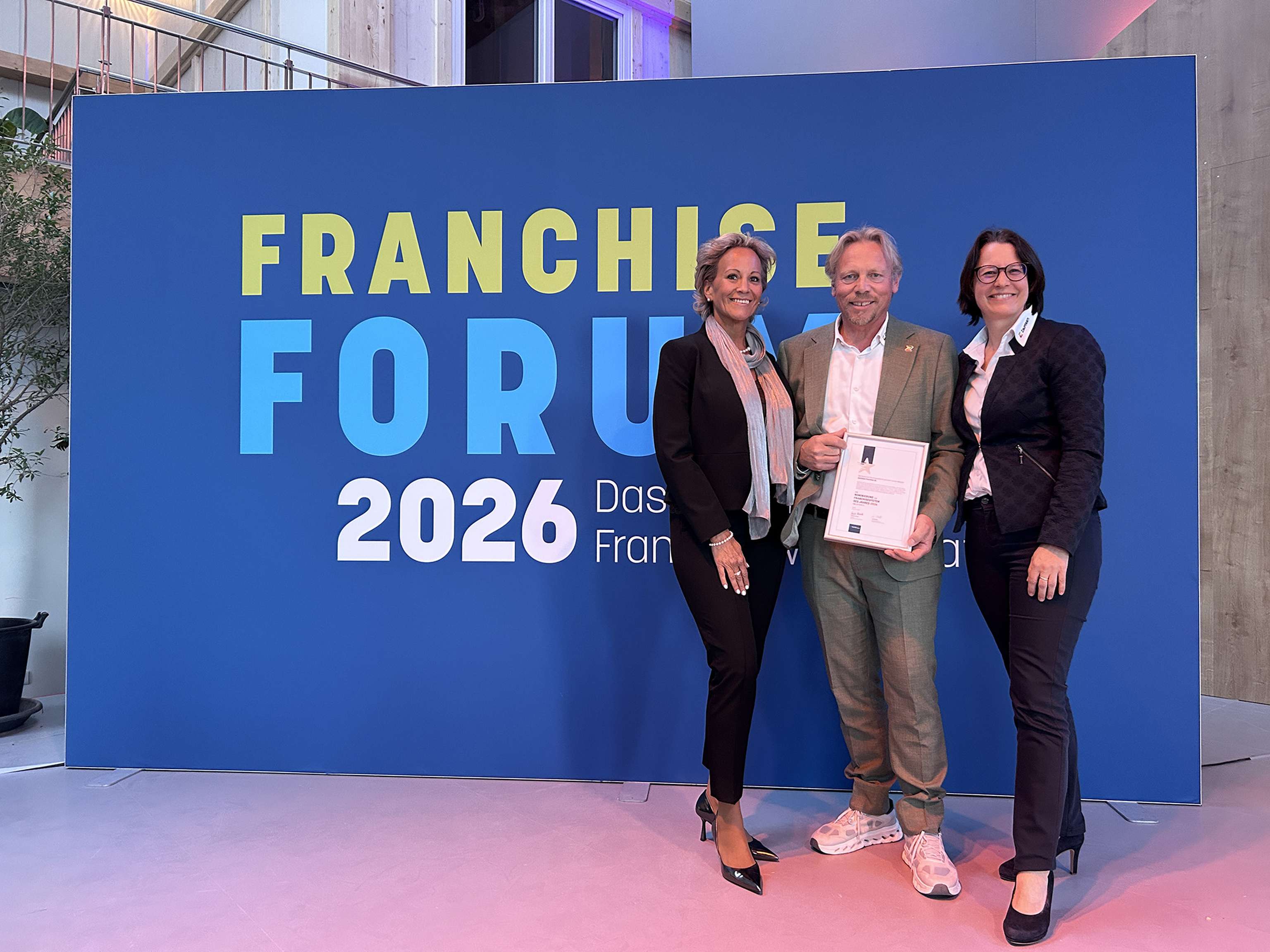 Drei Personen stehen vor einem blauen Schild mit der Aufschrift "Franchise Forum 2026", eine Person hält eine Urkunde in der Hand.