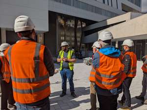 A group of people wearing orange safety vests and white hard hats listen to a speaker at a construction site.