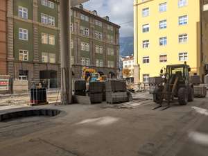 Urban construction site with machinery, stacks of materials, and surrounding buildings under a partly cloudy sky.