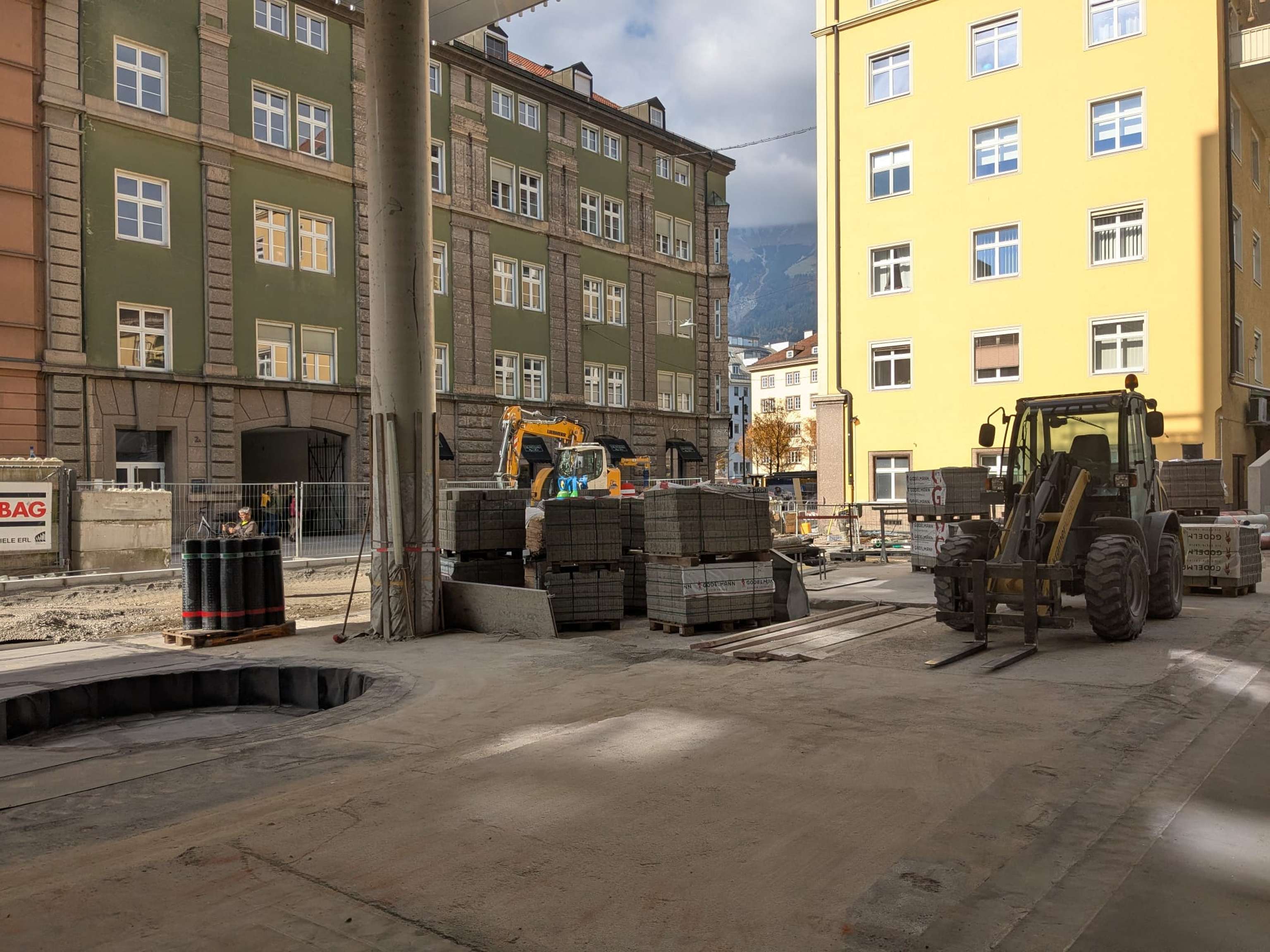 Urban construction site with machinery, stacks of materials, and surrounding buildings under a partly cloudy sky.