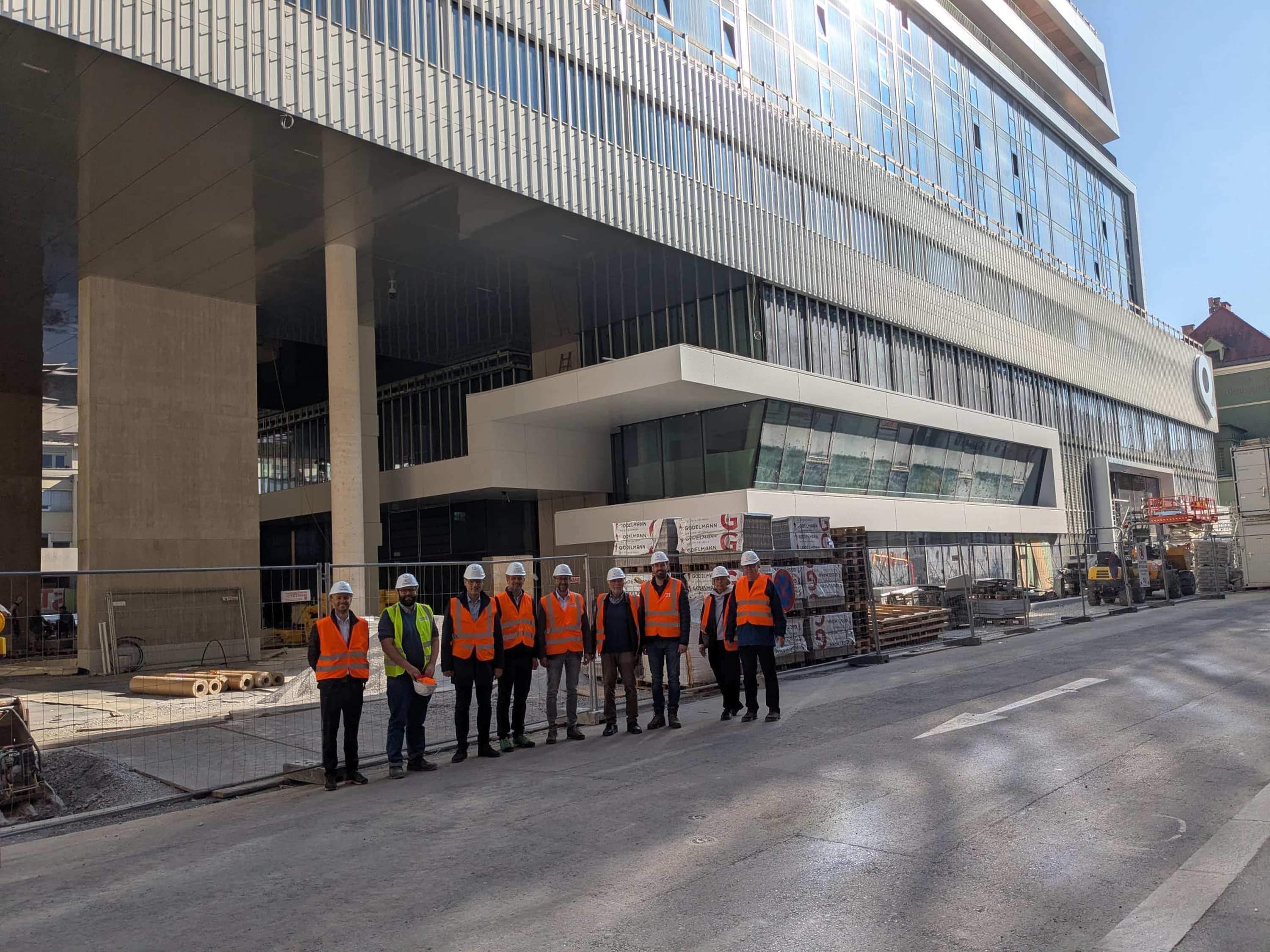 A group of construction workers in safety vests and helmets stand in front of a modern building under construction on a sunny day.