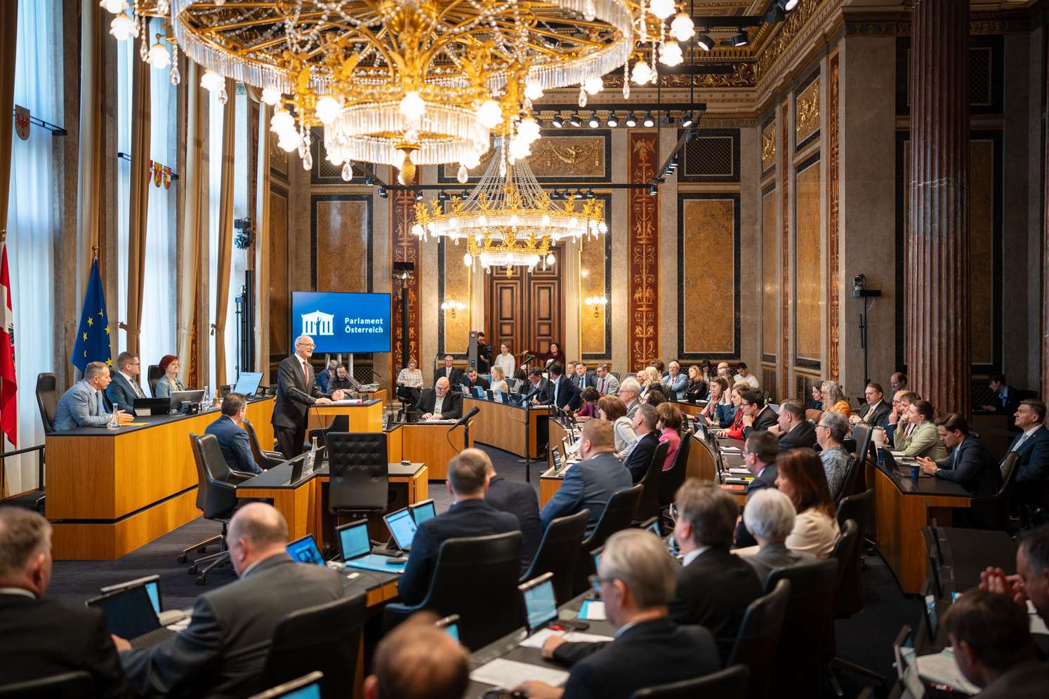 Versammlung in einem opulenten Saal mit Kronleuchtern und Beamten an Tischen, während ein Redner am Pult präsentiert.