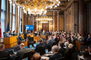 Versammlung in einem opulenten Saal mit Kronleuchtern und Beamten an Tischen, während ein Redner am Pult präsentiert.