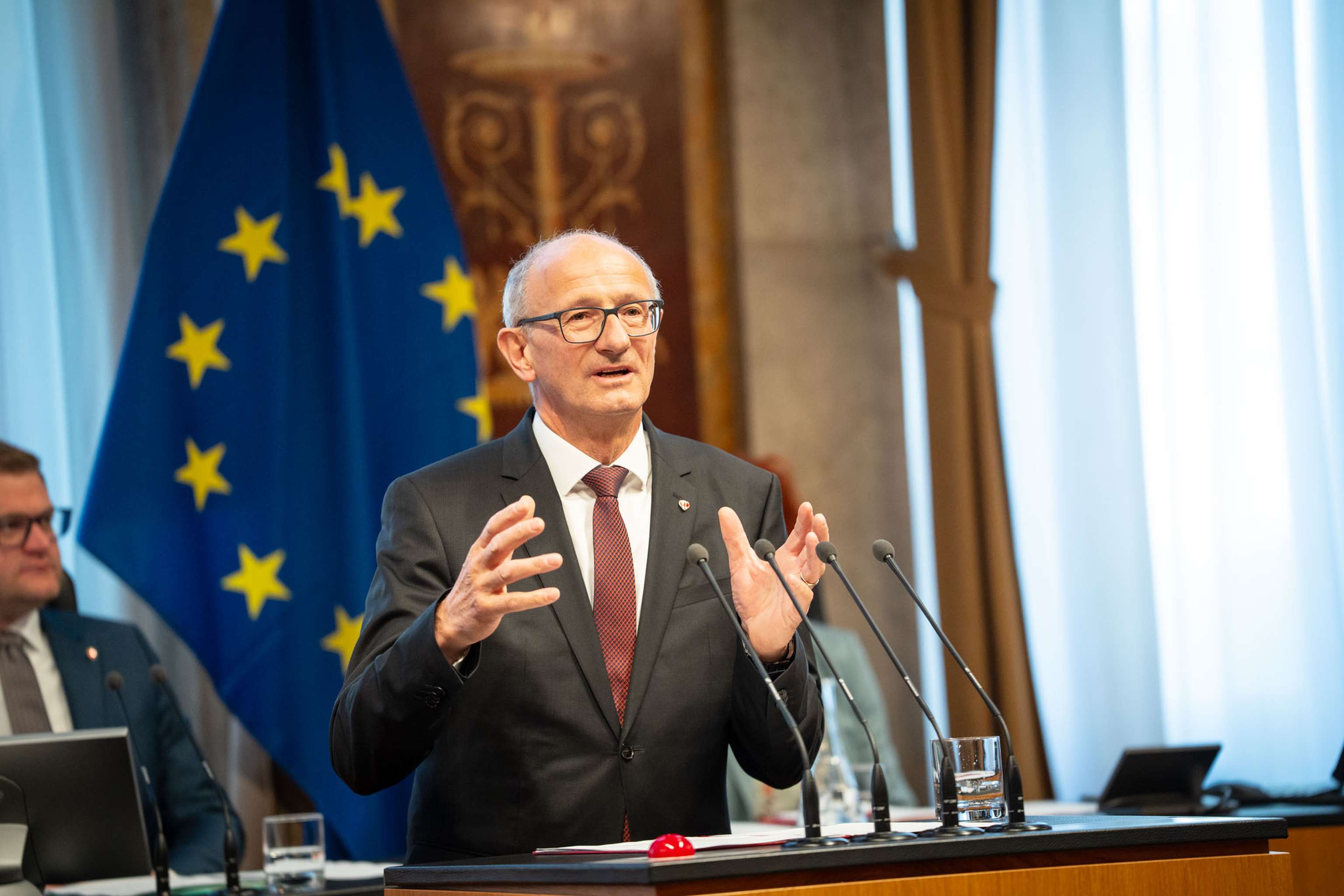 Ein Mann in Anzug hält eine Rede, hinter ihm eine EU-Flagge und ein Rednerpult.