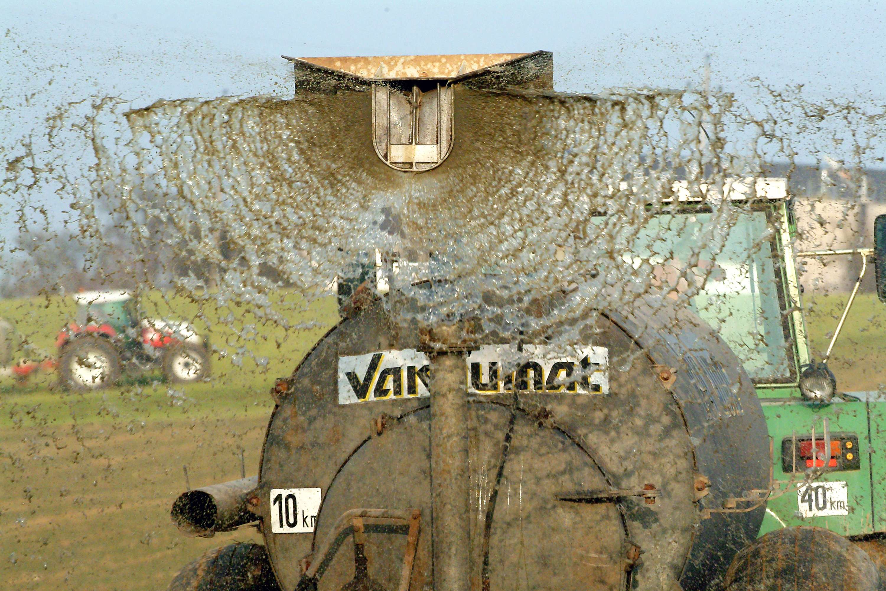 A tractor with a manure spreader sprays liquid manure across a field, with droplets fanning out widely. Another tractor is visible in the distance.
