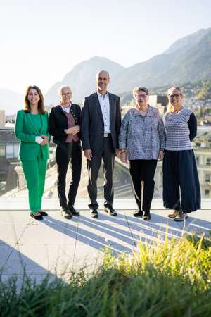 Fünf Personen posieren lächelnd auf einer Dachterrasse mit Bergpanorama im Hintergrund, in professioneller Kleidung.
