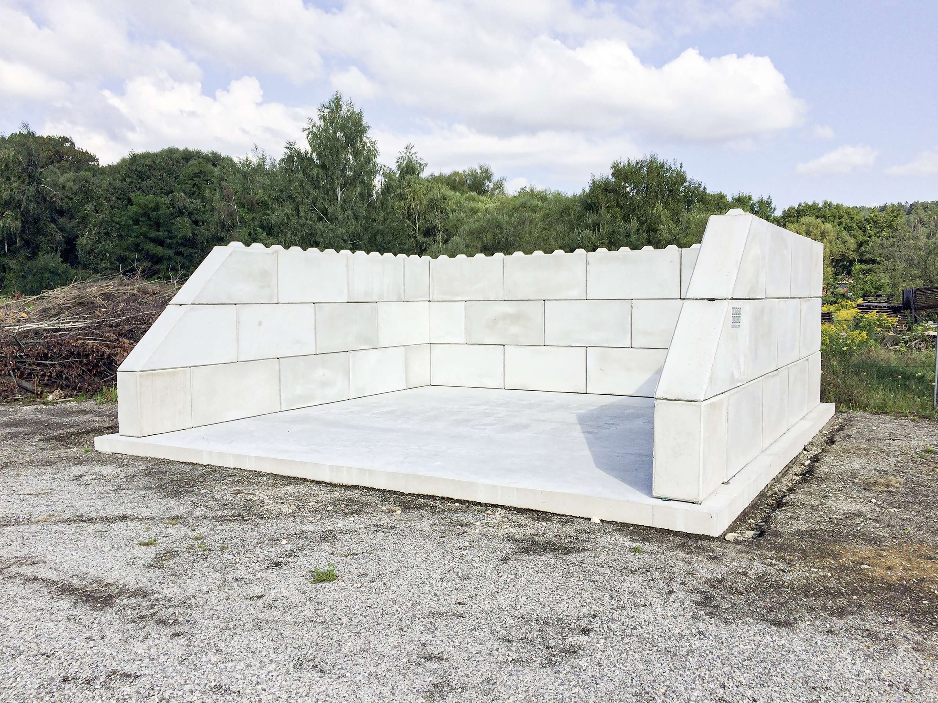 Concrete structure with tall walls on three sides, resembling an oversized open box, set on a gravel surface with trees and cloudy sky in the background.