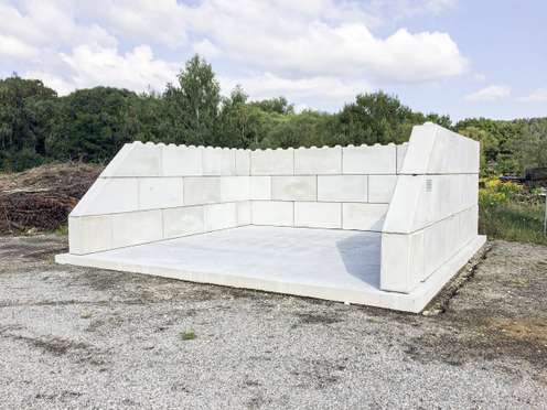 Concrete structure with tall walls on three sides, resembling an oversized open box, set on a gravel surface with trees and cloudy sky in the background.