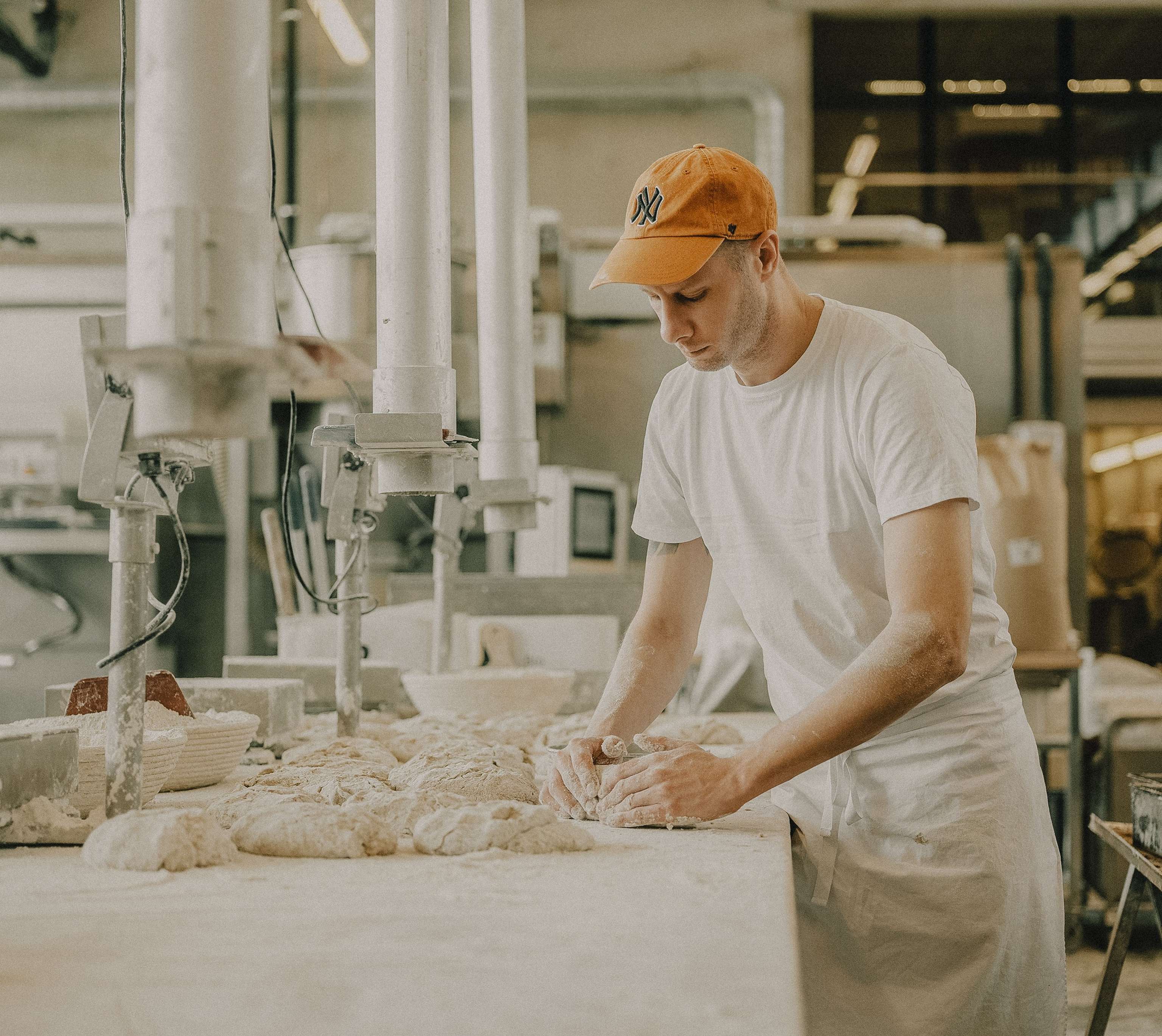Bäcker mit orangefarbener Kappe knetet Teig auf einer bemehlten Arbeitsfläche in einer gewerblichen Bäckerei.